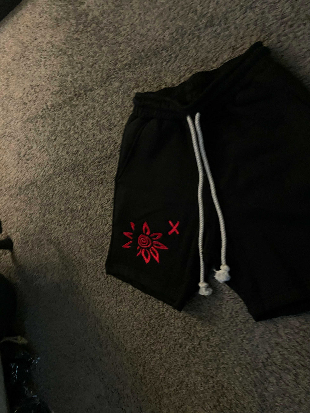Black shorts with red WISE Flower x embroidery and white drawstrings on carpet background