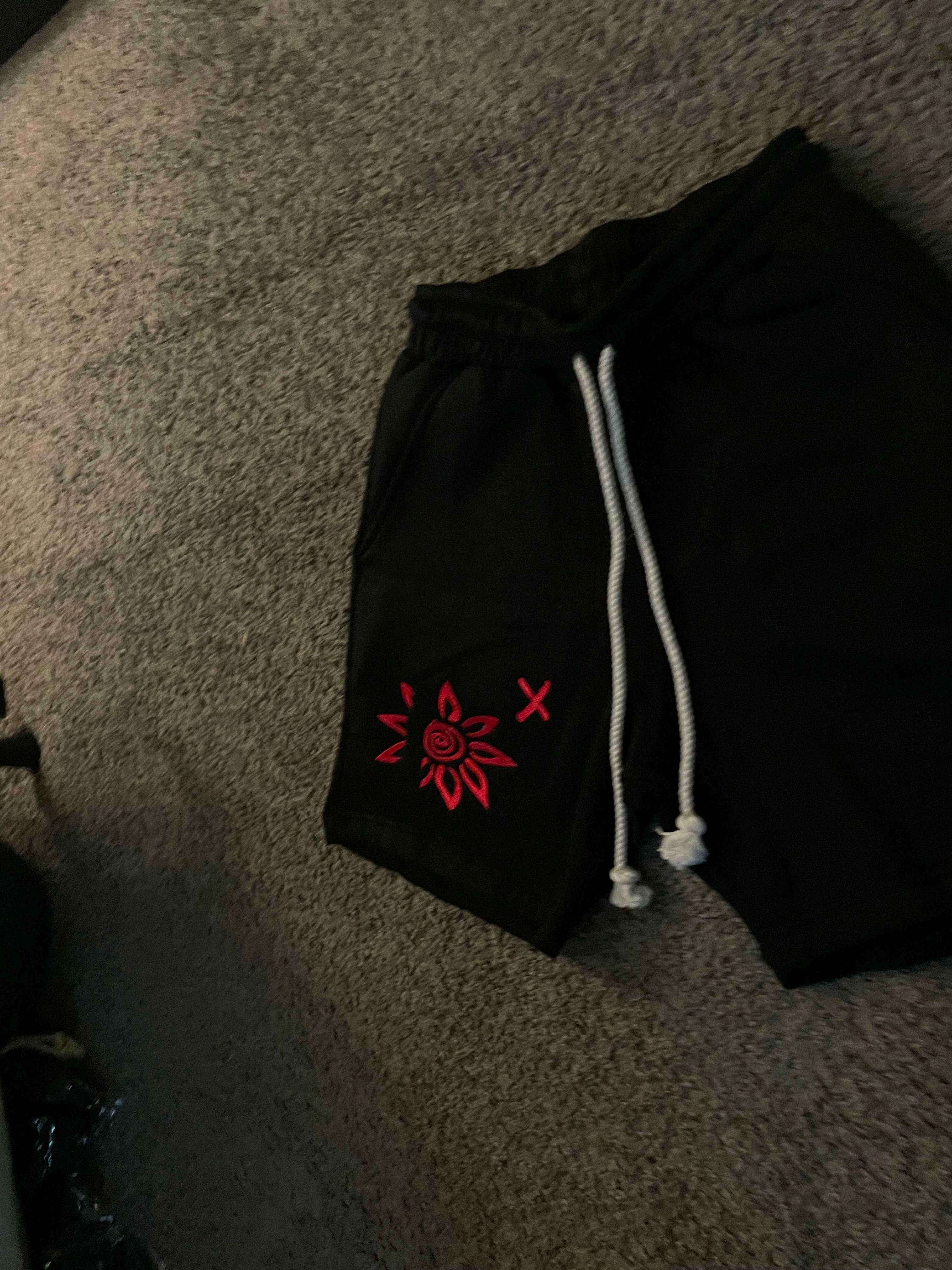 Black shorts with red WISE Flower x embroidery and white drawstrings on carpet background