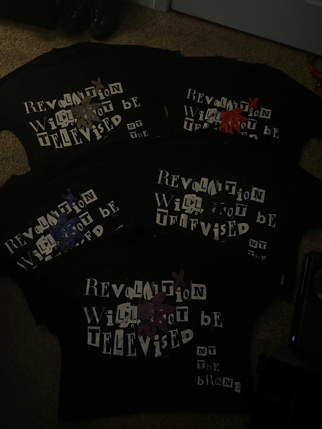 Set of black Revolution bundle t-shirts with colorful flower x designs and white text saying "Revolution will not be televised" arranged in a fan shape on the floor