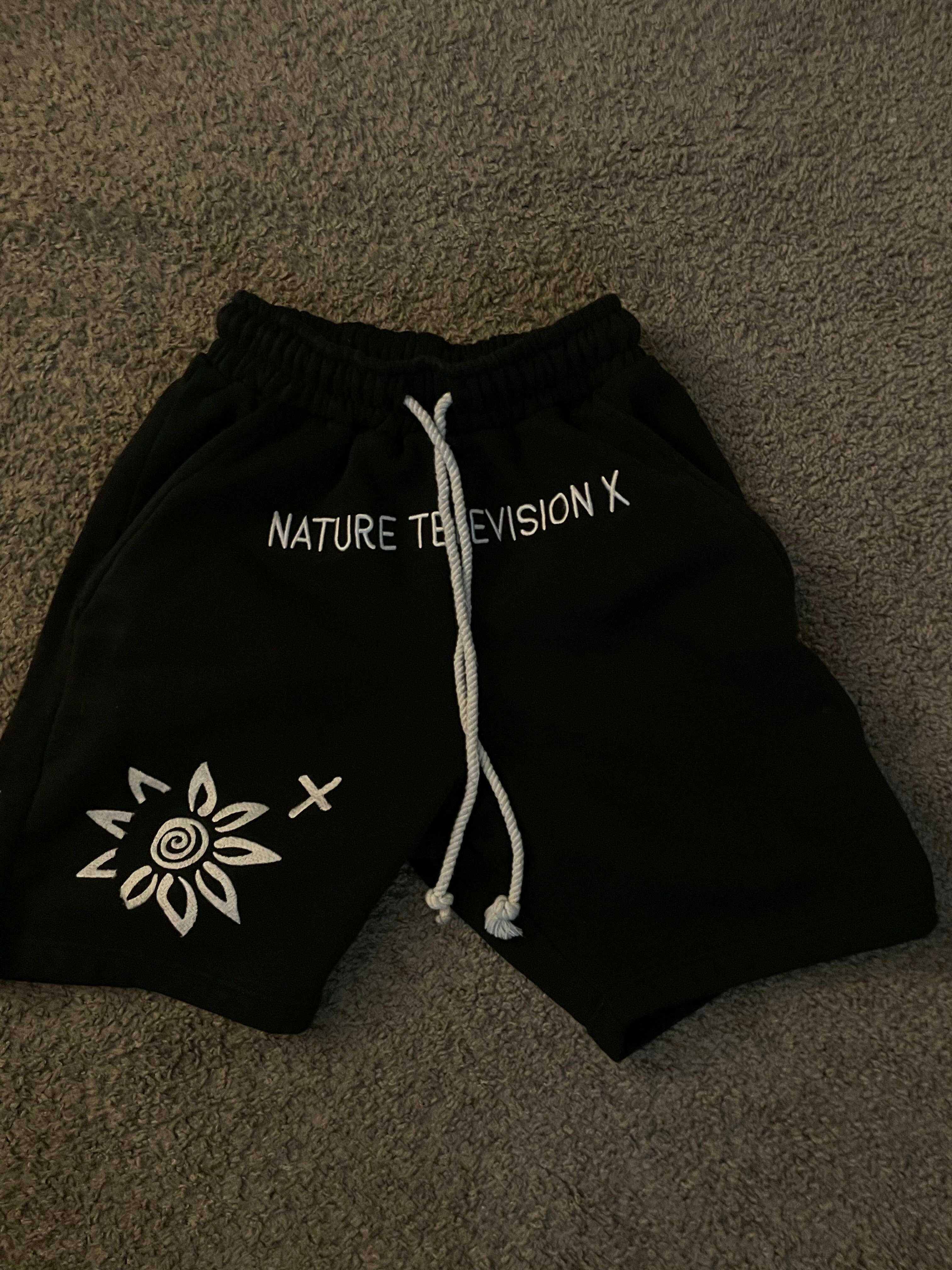 Black men's shorts with white "NATURE TELEVISION X" text and embroidered flower design, featuring a white drawstring, made of cotton and polyester, WISE FLOWER X brand
